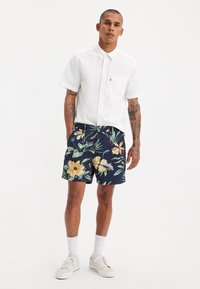 Cămașă albă cu mâneci scurte și nasturi, dotată cu un buzunar pe piept, asortată cu pantaloni scurți navy care au un imprimeu floral vibrant pe un fundal închis.