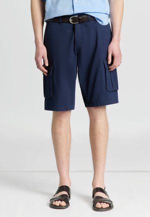 Uomo con pantaloncini cargo blu navy, cintura nera, camicia azzurro chiaro e sandali neri, in piedi su uno sfondo chiaro e uniforme.