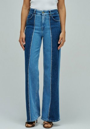 Femme portant un jean taille haute à jambes larges avec des panneaux verticaux bleu foncé et clair, un haut blanc et des sandales à talons foncées, debout devant un fond gris.