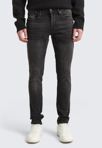 Jeans de denim preto com um corte ajustado, apresentando cinco bolsos, fecho de zip e um desbotado subtil. Combinados com ténis brancos.