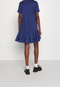 Mörkblå bomullsklänning med korta ärmar och volangkant. Bärs med svarta skor och vita strumpor, visad från en bakvy.