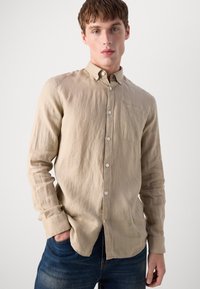 Camisa bege de manga longa com botão, em tecido texturizado, com bolso no peito e punho com um botão, combinada com jeans azul.