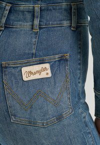 Klassiske blå denimjeans med en tydelig Wrangler-logo på baklommen, aksentuert med gule sømmer og et buet design.