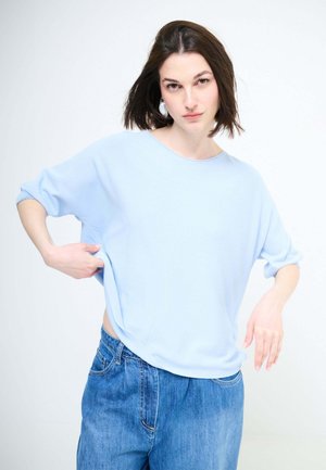 Giovane donna che sistema un maglione azzurro chiaro ampio, indossa jeans di denim blu, in piedi davanti a uno sfondo bianco semplice.