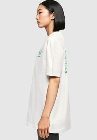 Weißes Baumwoll-T-Shirt, lässiger Schnitt, kurze Ärmel. Grüne Schrift und Grafiken vorne und hinten. Glatte Textur, runder Halsausschnitt.