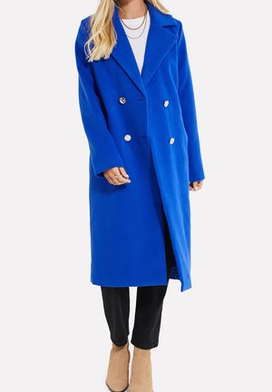 Femme portant un manteau long bleu vif à double boutonnage, un haut blanc, un pantalon noir, des bottines marron clair et plusieurs colliers en or superposés.