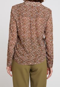 Brun blommig blus med långa ärmar, prydd med vita och rosa blommor på en texturerad bakgrund, tillsammans med olivgröna byxor.