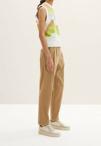 Szydełkowy bezrękawnik w zielone, pomarańczowe i fioletowe fale; zestawiony z beżowymi, plisowanymi spodniami o prostym kroju i jasnymi trampkami.