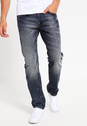 Mann trägt ausgewaschene schwarze Slim-Fit-Jeans und weiße Sneaker, Hand in der Tasche, geht vor weißem Hintergrund.