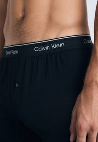 Boxeri negri din bumbac cu deschidere la buton, având un logo Calvin Klein în contrast pe banda elastică. Textură netedă.