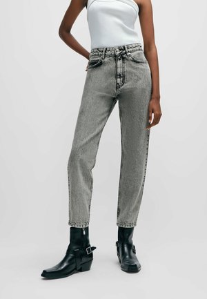 Personne portant un jean gris clair délavé, des bottines noires à boucles et un haut blanc sans manches sur un fond clair uni.