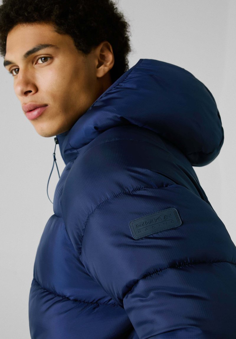 Donkerblauwe gewatteerde jas met capuchon, met een gestructureerde afwerking en een merkpatch op de linkerschouder.