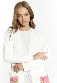 Cardigan bianco a maglia con maniche lunghe, dotato di tasche laterali con motivi geometrici rosa e gialli e dettagli con frange.