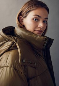 Brun polstret jakke med høj krave, synlige knapper og et struktureret stof. Designet inkluderer en lynlås lukning og en glat overflade.