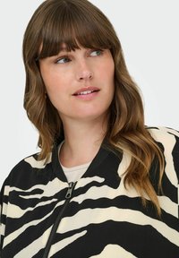Crna i krem prugasta bomber jakna sa zipom sprijeda, rebrastim ovratnikom i teksturiranim materijalom. Model ima valovitu kosu.