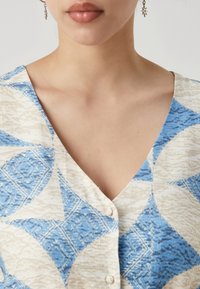 Vêtement en V avec un motif géométrique bleu et crémé, mettant en avant des formes de diamants et une texture. Des boutons sur le devant rehaussent le design.