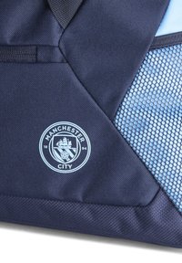 Bolsa de color azul marino con un panel de malla, logo del Manchester City en azul claro y tejido texturizado. Diseño geométrico con costuras reforzadas.