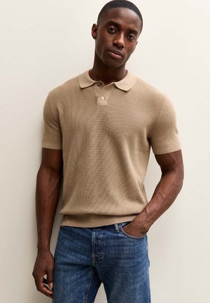 Homme debout, une main dans la poche, portant un polo en maille beige à manches courtes et un jean bleu, sur un fond clair uni.