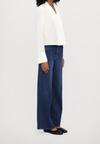 Blusa blanca de manga larga con escote en V y puños anchos, combinada con unos jeans de mezclilla oscuros de pierna ancha y cintura alta. Unos zapatos bajos negros completan el look.