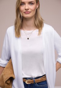 Young woman with blonde hair wears white cardigan over white shirt, blue jeans with tan belt, layered gold necklaces, holding tan jacket.