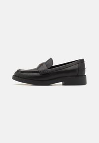 Black leather loafers with a stitched trim, flat rubber sole, and a decorative strap across the vamp; smooth texture and minimal design.