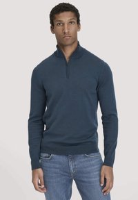 Dunkelblauer Zip-Up-Pullover mit Stehkragen, langen Ärmeln, gerippten Bündchen und Saum, aus einem glatten, weichen Material gefertigt. Über Jeans getragen.