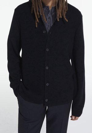 Persona con lunghi dreadlocks che indossa un cardigan nero abbottonato sopra una camicia scura a motivo e pantaloni neri, in piedi con le mani in tasca.