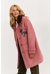 Ružový kordúrový kabát s hnedým kožušinovým golierom, zdobený výšivkou húb a prednými vreckami, nosený cez nohavice s leopardím vzorom.