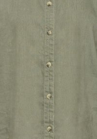 Chemise verte boutonnée avec un tissu lisse et léger. Elle comporte six boutons ronds de couleur claire et des coutures visibles à l'avant.