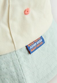 Gros plan sur une casquette beige et vert clair avec un trou de ventilation cousu et une étiquette bleue portant l'inscription « MAKE WAVES » en texte orange et blanc.
