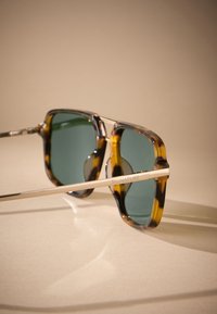 Lunettes de soleil à monture écaille de tortue avec des verres verts, une forme carrée, et des accents métalliques dorés sur les branches portant le mot gravé "SIGNATURE".
