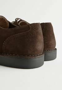Zapatos de ante marrón con tacones redondeados, acentos cosidos y una suela de goma negra, con una superficie texturizada y un acabado pulido.