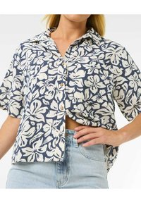 Kurzarmhemd aus marineblauem Leinen mit cremefarbenem Blumenmuster, durchgehende Knopfleiste und einer Tasche auf der linken Seite, kombiniert mit hellblauen Jeans.