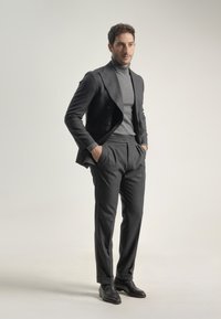 Hombre con traje gris oscuro, suéter de cuello alto gris, zapatos negros, de pie con las manos en los bolsillos frente a un fondo claro y sencillo.