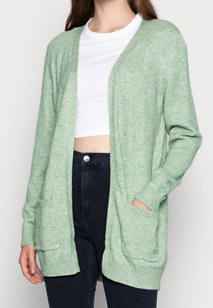 Woman wearing a light green open-front cardigan with front pockets over a white cropped top and black high-waisted jeans.