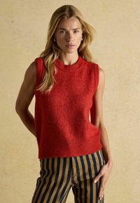 Roter ärmelloser Pullover aus weichem, strukturiertem Material mit geripptem Kragen und Saum. Kombiniert mit navy- und braun gestreiften Hosen mit vertikalen Streifen.