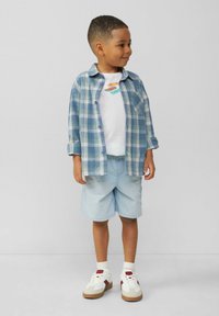 Jongen in een blauw-groen geruit shirt over een wit T-shirt, lichte blauwe shorts en witte sneakers met rode accenten.