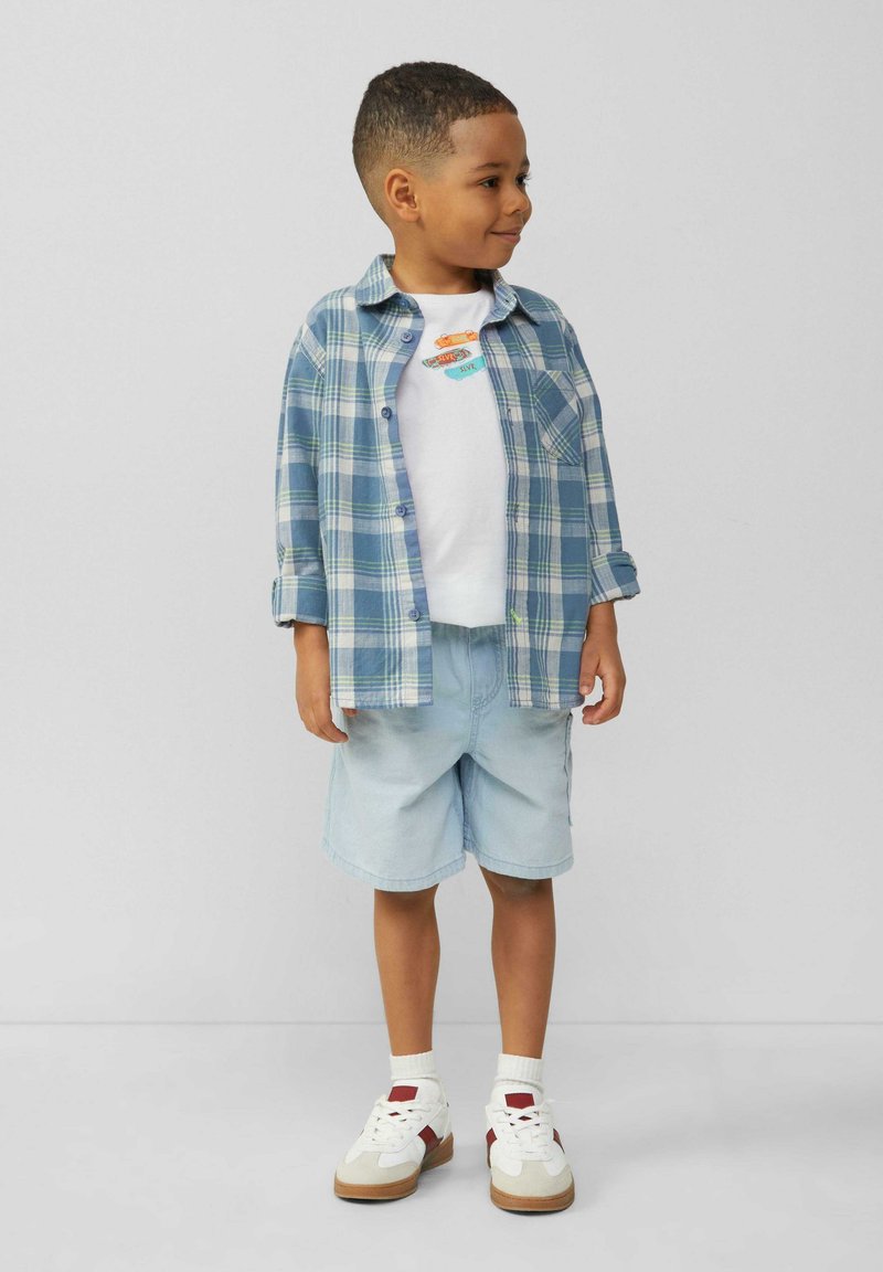 Jongen in een blauw-groen geruit shirt over een wit T-shirt, lichte blauwe shorts en witte sneakers met rode accenten.