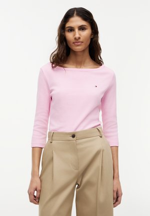 Kobieta w jasnoróżowej bluzce z rękawami do łokcia i beżowych spodniach z wysokim stanem, stojąca na tle jednolitego białego tła.