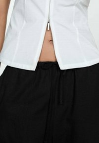 Haut blanc à manches courtes avec une fermeture éclair au centre, associé à un pantalon noir avec cordon de serrage. Les tissus semblent lisses et légers.