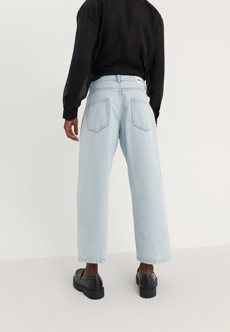 Jean large bleu clair à taille haute, avec poches arrière et ourlet cousu. Associé à une chemise noire transparente à manches longues et des chaussures noires.