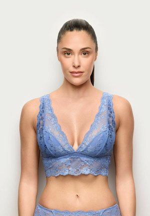 Frau trägt ein blaues Spitzen-Bralette und passende Unterwäsche, steht vor einem schlichten hellen Hintergrund, mit zurückgebundenen Haaren.