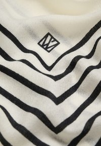 Kreemi kangas mustade ebakorrapäraste zikk-zakk triipudega ja väike must geomeetriline logo keset voldi lähedal.