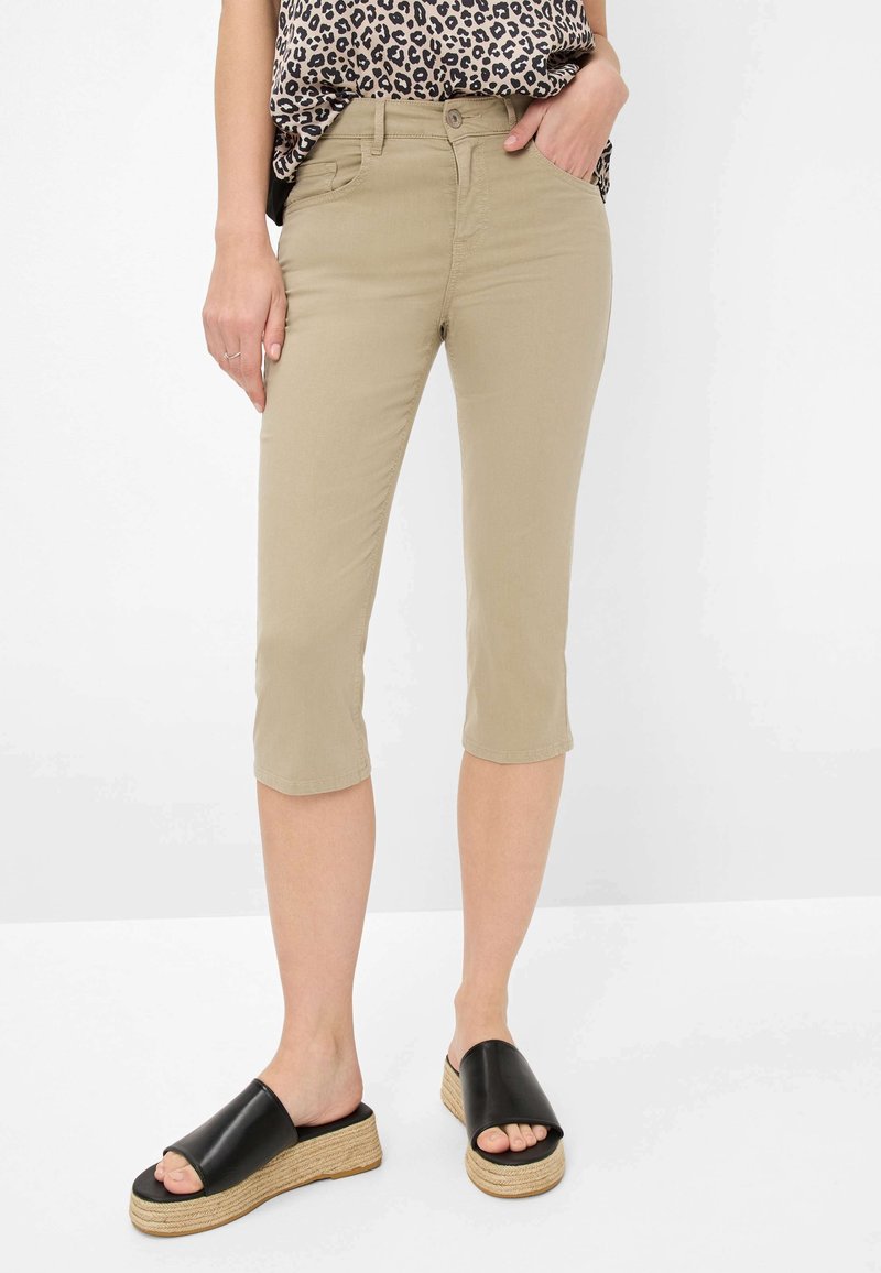 Femme portant un pantalon beige court, un haut à imprimé léopard et des sandales compensées noires avec plateforme tissée, debout sur fond blanc.
