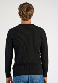 Zwarte gebreide trui met een gestructureerd ribpatroon, ronde halslijn en aansluitende manchetten. Gecombineerd met blauwe denim jeans.