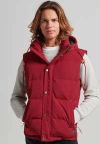 Doudoune rouge avec capuche, dotée d'une fermeture éclair, de boutons-pression, de deux poches latérales et de cordons de serrage élastiques. L'intérieur présente une doublure contrastante.