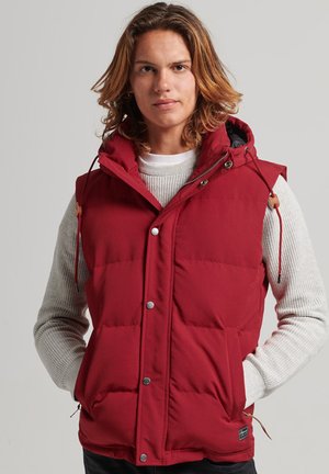 Red puffer vest with a hood, featuring a zip closure, snap buttons, two side pockets, and elastic drawstrings. The inside has a contrasting lining.
