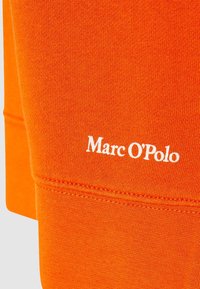 Tela de algodón naranja con textura de punto visible, con la marca blanca "Marc O’Polo". Detalles de costura destacados con costuras en contraste.
