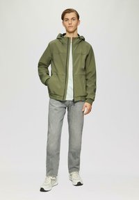 Olivegrüne Kapuzenjacke mit Vorderreißverschluss, seitlichen Taschen und figurnaher Passform, kombiniert mit einem hellgrünen Shirt und grauen Jeans, getragen mit Sneakers.