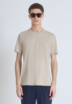 Camiseta de manga corta beige con cuello redondo y detalle de logo en relieve. La tela parece suave y ligera, adecuada para un uso casual.